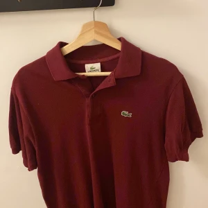 Vinröd pikétröja från Lacoste - Säljer en stilren vinröd pikétröja från Lacoste med klassisk krage och knappslå. Tröjan har det ikoniska gröna krokodilemblem på bröstet. Perfekt för en avslappnad men snygg look.