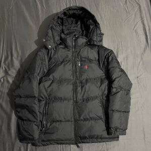 Svart Polo dunjacka med huva - Säljer en svart dunjacka från Polo Ralph Lauren med huva och dragkedja. Jackan har en röd logotyp på bröstet och praktiska fickor med dragkedja. Perfekt för kalla dagar. Storlek L passar mer som M