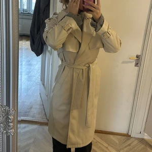 Beige trenchcoat från JUSTFAB - Snygg beige trenchcoat från JUSTFAB med klassisk design i läderliknande material. Jackan har ett bälte i midjan och långa ärmar med spännen vid ärmsluten. Perfekt för höst och vår!