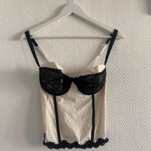 Vit korsett med svarta spetsdetaljer - Elegant vit korsett med svarta spetsdetaljer och justerbara axelband. Den har en feminin design med spets över bysten och längs nederkanten. Perfekt för en stilfull look.