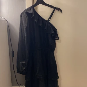 Svart klänning från Lindex - Snygg svart klänning från Lindex med en unik oneshoulder-design och volangdetaljer. Klänningen har långa ärmar och är perfekt för speciella tillfällen. Den är tillverkad i ett lätt och bekvämt material.