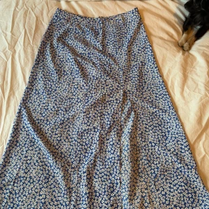 Blommig långkjol i blått och vitt - Charmig långkjol med blommigt mönster i blått och vitt. Perfekt för en somrig look med sitt lätta och luftiga material. Kjolen har en elegant passform som ger en fin siluett.