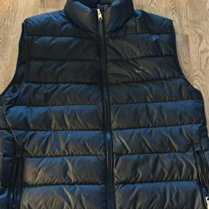 Svart dunväst från GANT - Snygg svart dunväst från GANT med dragkedja framtill. Perfekt för kyligare dagar och ger en stilren look. Västen har en quiltad design som ger extra värme och komfort. hör av er om ni har frågor