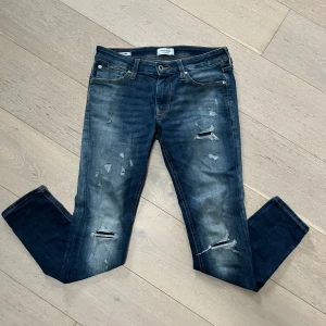 Jack and Jones jeans Mörkblå jeans med slitningar - Snygga mörkblå jeans med coola slitningar och en klassisk femficksdesign. De har en dragkedja och knapp framtill. Perfekta för en avslappnad stil med en touch av edge.