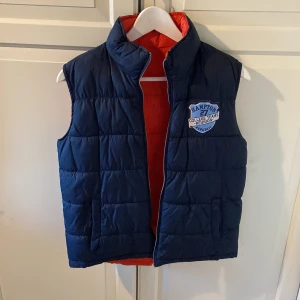 Vändbar dunväst i blått och orange - Snygg vändbar dunväst från Hampton med en sida i marinblått och den andra i orange. Västen har ett emblem på bröstet och dragkedja framtill. Perfekt för att variera din stil beroende på humör eller tillfälle.