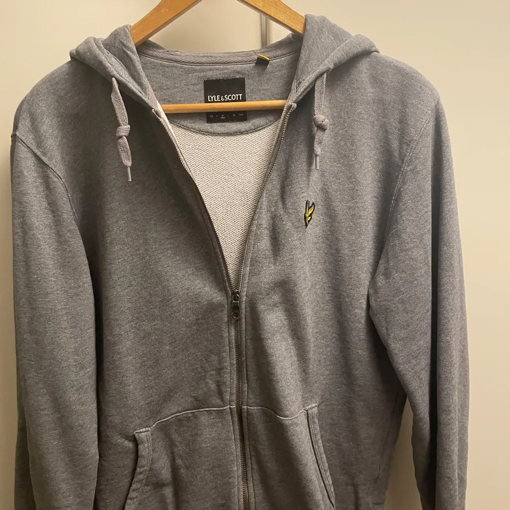 Säljer en stilren grå hoodie från Lyle & Scott med dragkedja och huva. Den har ett broderat gult emblem på bröstet och praktiska fickor framtill. Perfekt för en avslappnad look.. Neuletakit & Villapaidat.
