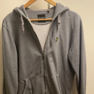 Grå hoodie från Lyle & Scott - Säljer en stilren grå hoodie från Lyle & Scott med dragkedja och huva. Den har ett broderat gult emblem på bröstet och praktiska fickor framtill. Perfekt för en avslappnad look.