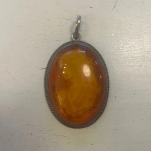 Ovalt hängsmycke med porträtt i silver och bärnsten - Elegant ovalt hängsmycke i silver med ett detaljerat porträtt i en varm bärnsten. Smycket har en dekorativ kant och en ögla för kedja. Perfekt för att ge en vintagekänsla till din stil. 5,5cm