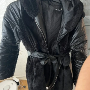 Svart pälsjacka med bälte - Snygg svart pälsjacka från Chich med quiltade detaljer och bälte i midjan. Jackan har en mysig huva och dragkedja framtill. Perfekt för kyliga dagar när du vill hålla stilen.