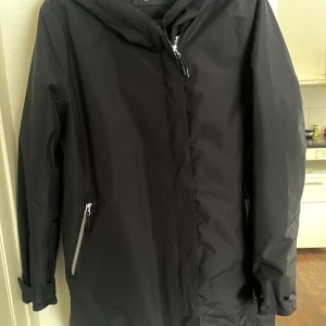 Svart parkajacka med dragkedja - Säljer en stilren svart parkajacka med huva och dragkedja framtill. Jackan har långa ärmar och två praktiska fickor med dragkedja. Perfekt för kyligare dagar. En jacka som är allround vatten avvisande från Everest.