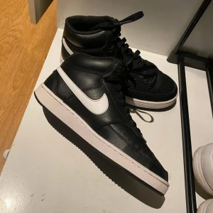 Svarta Nike sneakers med vit sula - Snygga svarta Nike sneakers med vit sula och vit swoosh-logga. Skorna har en hög modell med snörning och är tillverkade i skinn. Perfekta för en stilren look. 