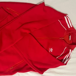Adidas Tröja - Säljer en fin röd träningsjacka från Adidas med klassiska vita ränder på axlarna. Storlek S och är i god skick.