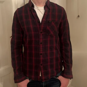 Röd och svart rutig skjorta från Lee - Snygg röd och svart rutig skjorta från Lee med långa ärmar och klassisk krage. Skjortan har knappar framtill och en bröstficka med Lee logga. Perfekt för en avslappnad stil.