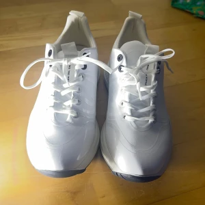 Vita Golfskor  från Duca del Cosma - Snygga vita golfskor från Duca del Cosma med snörning och en stilren design. Skorna har en svart sula med mönster för bra grepp på golfbanan och en diskret logga på sidan. Perfekta för en sportig och modern look när man spelar golf. Nypris: Ca 2000kr