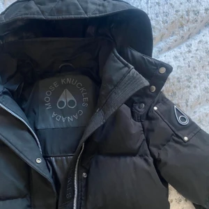 Svart dunjacka från Moose Knuckles - Säljer en svart dunjacka från Moose Knuckles med huva, jätte fint skick då den knappt är använd, enda är att dragkedjan vid rumpan åkt ur lite i mitten men man kan enkelt trycka tillbaka den om man vill, syns inte och som sagt enkelt att fixa