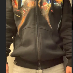 Svart hoodie med färgglatt tryck - Snygg svart hoodie med dragkedja och ett färgglatt tryck på bröstet. Den har långa ärmar och en justerbar huva med snören finns ingen skador 