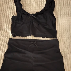 Svart pyjamas set från SHEIN - Säljer en svart ribbad linnetopp med volangdetaljer och en liten rosett framtill. Toppen är croppad och har breda axelband. Perfekt för en stilren och bekväm look.