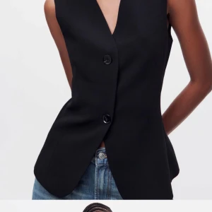 Svart väst med knappar - Elegant svart väst med djup v-ringning och stora knappar framtill. Västen har en stilren design med ett bälte baktill för en justerbar passform. Perfekt för en chic och modern look.
