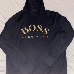 Svart hoodie från Hugo Boss - Snygg svart hoodie från Hugo Boss med guldtryck på bröstet. Den har en klassisk design med huva och dragsko. Perfekt för en stilren och bekväm look.