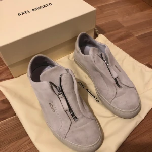 Beiga sneakers från Axel Arigato - Snygga grå sneakers från Axel Arigato i mocka med dragkedja framtill. Tillverkade i Portugal och kommer med originalförpackning. Perfekta för en stilren look. Storlek 41
