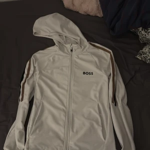 BOSS Kofta  - Säljer en stilren vit huvtröja från Boss med dragkedja och bruna detaljer på ärmarna. Som ny köpt för 2400 ( den finns inte längre i marknaden)