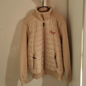 Beige quiltad jacka från Hugo boss - Säljer en beige quiltad jacka från HUGO med dragkedja och broderad logga i rött på bröstet. Jackan har långa ärmar och två fickor med dragkedja framtill. Perfekt för en stilren look! Har aldrig använt!!! 