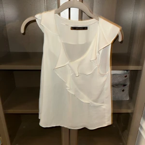 Vit blus från Zara - Elegant vit blus från Zara med volangdetaljer framtill. Blusen är ärmlös och har en mjuk, flytande design som ger en stilren look. Perfekt för att klä upp eller ner beroende på tillfälle.