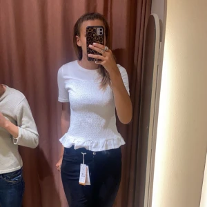 Smockad topp - Säljer en söt vit topp med korta ärmar och volangdetalj vid nederkanten. Toppen har en smockad textur framtill som ger en fin passform. Perfekt för både vardag och fest!