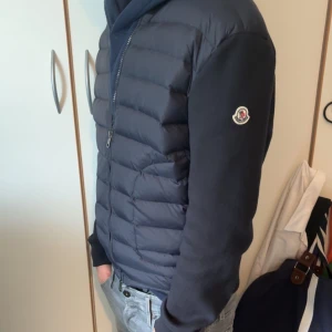 Mörkblå pufferjacka från Moncler - Säljer en snygg mörkblå pufferjacka från Moncler. Jackan har en dragkedja framtill och långa ärmar. Perfekt för kyligare dagar med sin quiltade design som håller värmen. Moncler-loggan syns på ärmen för en stilren look.