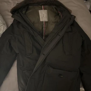 Mörkgrön parkajacka från Fortezza - Säljer en stilren mörkgrön parkajacka från Fortezza. Jackan har en quiltad insida och en mysig krage. Den är utrustad med flera praktiska fickor och en dragkedja framtill. Perfekt för kyligare dagar.