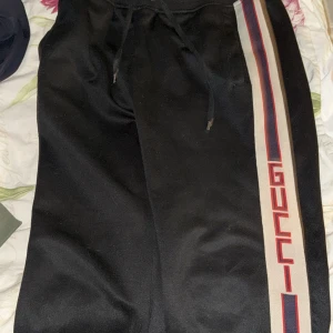 Svarta mjukisbyxor från Gucci - Snygga svarta mjukisbyxor från Gucci med röd och vit randig detalj längs sidan och logotyp. Byxorna har snörning i midjan och ribbade muddar vid bensluten. Perfekta för en avslappnad stil med en lyxig touch.