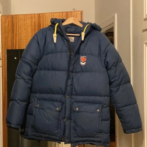Blå dunjacka från Fjällräven - Säljer en blå dunjacka från Fjällräven med dragkedja och knappar. Jackan har långa ärmar och praktiska fickor framtill. Perfekt för kalla dagar med sin varma vaddering och justerbara huva med gula detaljer.