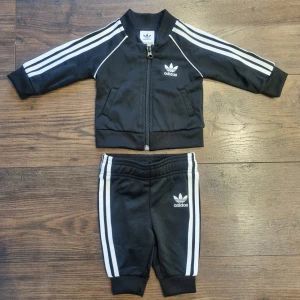 Svart tracksuit träningsset för bebis från Adidas. Ny - Ny svart tracksuit träningsset för bebis från Adidas med de klassiska tre vita ränderna längs ärmar och ben. Setet består av en jacka med dragkedja och matchande byxor. Perfekt för små sportfantaster! Storlek 62