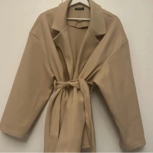 Beige kappa från Vero Moda - Snygg beige kappa från Vero Moda med knytband i midjan. Den har en klassisk krage och långa ärmar, perfekt för höst och vår. Stilren design som passar till många olika outfits.