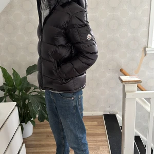 Svart Moncler maya - Säljer en snygg svart pufferjacka från Moncler. Jackan har en glansig finish och är utrustad med en dragkedja framtill. Den har en praktisk huva och är perfekt för kyligare dagar. Jackan har en normal passform och är långärmad.