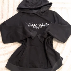 Svart hoodie med tribaltryck - Snygg svart hoodie med ett coolt vitt tribaltryck på bröstet. Perfekt för en avslappnad stil. Hoodien har en stor ficka framtill och en bekväm huva.