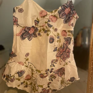 Blommig korsettopp - Säljer en vacker blommig korsettopp med ett mönster i rosa och blå toner. Toppen har en oneshoulder-design och volangdetaljer nedtill. Perfekt för att ge en feminin touch till din outfit . storlek S och M