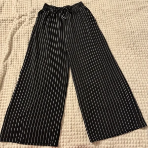Svarta randiga byxor - Snygga svarta byxor med vita ränder och en loose passform. De har en bekväm resår i midjan och vida ben som ger en avslappnad look. Perfekta för en stilren outfit. Om ni vill se den på, skriv gärna!