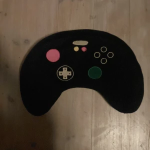 Svart kudde formad som en spelkontroll - En unik svart kudde formad som en spelkontroll med färgglada knappar i rosa, grön och gul. Perfekt för spelentusiaster som vill ha en rolig och annorlunda inredningsdetalj.