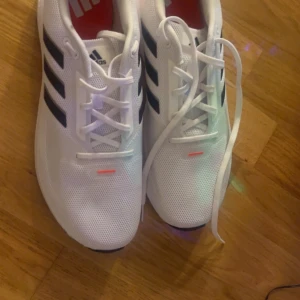 Vita Adidas sneakers med svarta ränder - Snygga vita Adidas sneakers med klassiska svarta ränder och snörning. Perfekta för en sportig look. Skorna har en bekväm sula och är tillverkade i syntetmaterial.