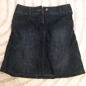 Mörkblå jeanskjol - Snygg mörkblå jeanskjol med klassisk femficksdesign och knappstängning fram. Kjolen har dekorativa sömmar och en broderad detalj på bakfickan. Perfekt för en avslappnad stil.