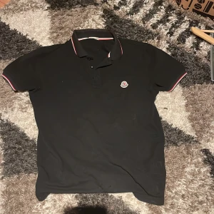 Svart pikétröja från Moncler - Snygg svart pikétröja från Moncler med klassisk krage och broderad logotyp på bröstet. Tröjan har detaljer i rött och vitt på kragen och ärmsluten. Sliten men de bara vid kragen säljer billigt då därför.