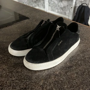 Svarta sneakers med dragkedja - Snygga svarta sneakers med dragkedja framtill. Skorna har en vit sula och är tillverkade i mocka och skinn, vilket ger en stilren look. Perfekta för en avslappnad stil.