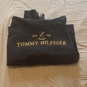 Svart hoodie från Tommy Hilfiger - Säljer en svart hoodie från Tommy Hilfiger med guldfärgad logotyp och text 'EST. 1985' på framsidan. Tröjan har en klassisk känguruficka och ribbade muddar vid ärmslut. Perfekt för en avslappnad stil.