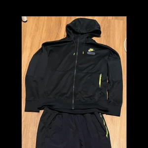 Svart nike airmax dress - Snygg svart hoodie och matchande byxor från Nike Air Max med gula detaljer. Hoodien har dragkedja och långa ärmar, perfekt för träning eller vardagsbruk. Byxorna har elastisk midja och sidofickor med dragkedja.