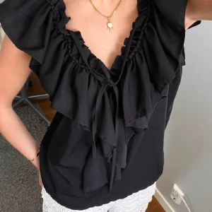 Svart blus med volangdetaljer - Snygg svart blus med volangdetaljer och knytning framtill. Perfekt för en stilren look. Blusen har en lös passform och är ärmlös. Jätte snygg. Köpt här på plick men den kom tyvärr aldrig till andvändning därför säljer jag. Sista bilden är på baksidan av tröjan. Det är inte mina bilder. Kontakta gärna vid frågor eller intresse. Pris kan diskuteras💗😊