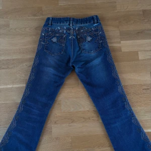 Blå bootcut jeans med broderi - Snygga blå bootcut jeans med detaljerat broderi längs sidorna och bakfickorna. Perfekta för en unik stil med en touch av vintage. De har en klassisk femficksdesign och en bekväm passform.