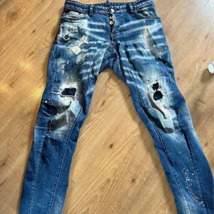 Blå jeans från Dsquared2 - Snygga blå jeans från Dsquared2 med slitna detaljer och färgstänk för en unik look. De har en knappgylf och klassisk femficksdesign. Perfekta för dig som vill ha en edgy stil.