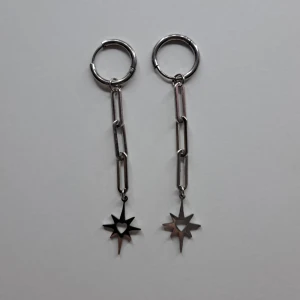Silvriga örhängen med stjärndetalj - Snygga silvriga örhängen med en unik design. De har en kedja med länkar och en hängande stjärna med ett utskuret hjärta i mitten. Perfekta för att ge en edgy touch till din stil. PRISET KAN DISKUTERAS