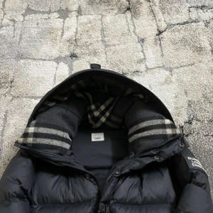 Svart dunjacka med rutigt foder - Säljer en stilren svart dunjacka från Burberry med ett snyggt rutigt foder. Jackan har en dragkedja och knappar framtill samt en mysig huva. Perfekt för kyliga dagar!